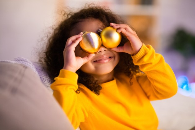 Little girl in yellow sweatshirt being silly and holding two yellow ornaments to her eyes. 