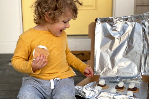 A toddler holding a s’more and looking at a homemade solar cooker.